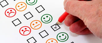 Mano che compila un questionario di soddisfazione segnando una casella accanto a una faccina sorridente verde, tra altre faccine con espressioni diverse.