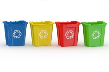 Immagine con quattro contenitori per la raccolta differenziata disposti in fila su sfondo chiaro. I bidoni sono di plastica e hanno tutti lo stesso design rettangolare con bordo superiore sporgente. Da sinistra a destra i colori sono: blu, giallo, rosso e verde. Su ciascun contenitore è presente il simbolo del riciclo bianco, ben visibile al centro. L’immagine è pulita, senza altri oggetti o elementi di distrazione, e comunica visivamente il concetto di separazione dei rifiuti.