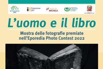 Locandina promozionale della mostra fotografica "L'uomo e il libro", dedicata alle fotografie premiate nell'Eporedia Photo Contest 2022. La grafica presenta, al centro, una fotografia in bianco e nero che ritrae un antico libro aperto sostenuto dalle mani segnate dal tempo di una persona, simbolo del rapporto tra l'uomo e la lettura. Nella parte superiore sono riportati i loghi della Rete Museale AMI, Fondazione CRT, Progetto Michela, Ecomuseo Anfiteatro Morenico di Ivrea e del Comune di San Giorgio Canavese. La mostra è visitabile dal 12 luglio al 18 ottobre 2026 presso il Museo Civico "Nòssi Ràis", in Via Campeggio 8, San Giorgio Canavese (TO), con apertura la domenica dalle 10:00 alle 12:00 e dalle 15:00 alle 18:00. In fondo alla locandina sono riportati i contatti dell'Ecomuseo AMI e i riferimenti ai canali social.